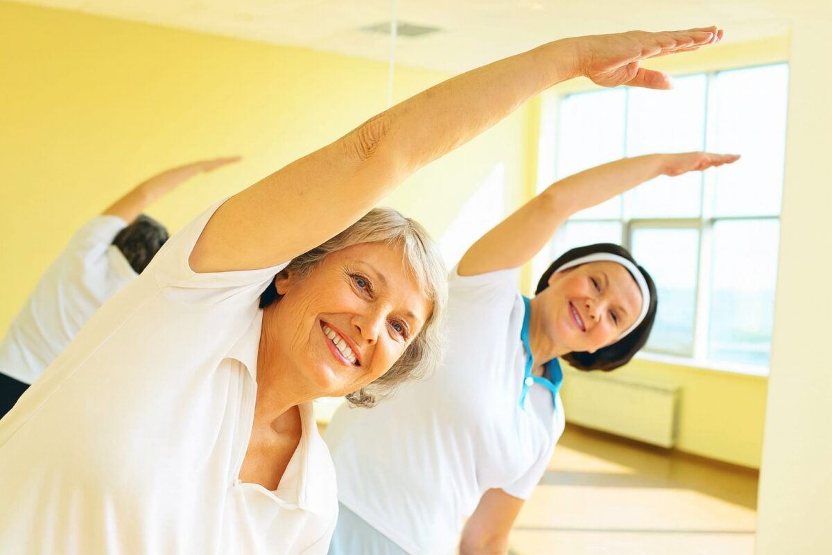 Older women stretching together in a sunlit fitness room, symbolizing active aging and occupational health support. Older women stretching together in a sunlit fitness room, symbolizing active aging and occupational health support.